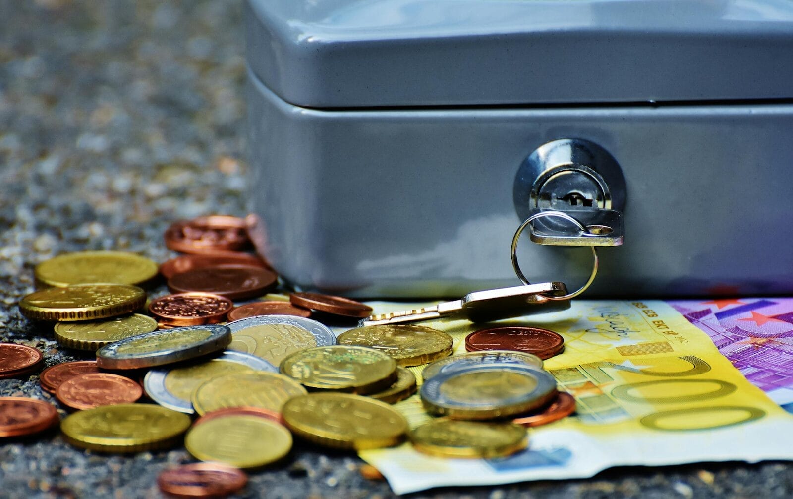 Gray safe deposit box with money strewn beside it. 